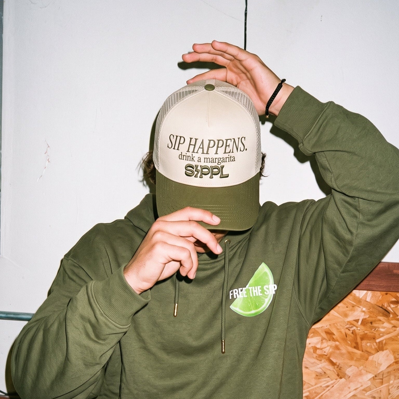 A person wearing an olive green hoodie and matching sweatpants sits indoors, holding a beige and green trucker hat over their face. The hat has the text 'SIP HAPPENS.