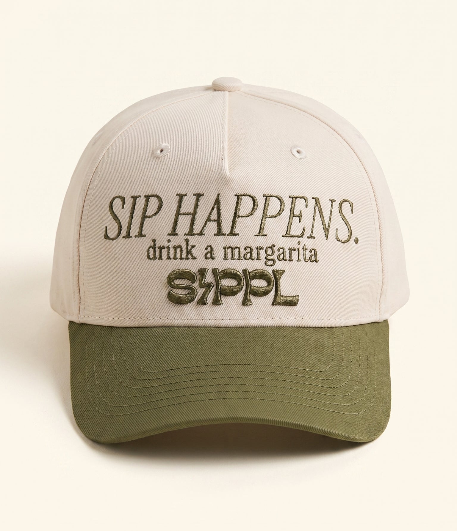 A structured baseball cap with a light beige crown and an olive green curved brim. The front of the cap features dark brown embroidered text that reads 'SIP HAPPENS.